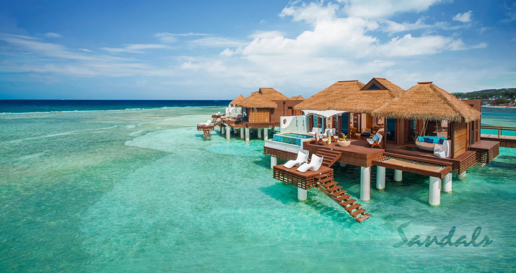 Thatched-roof overwater bungalows of a Royal Caribbean resort stretch out over a crystal clear sea, enhanced by a stunning blue skies in the background_